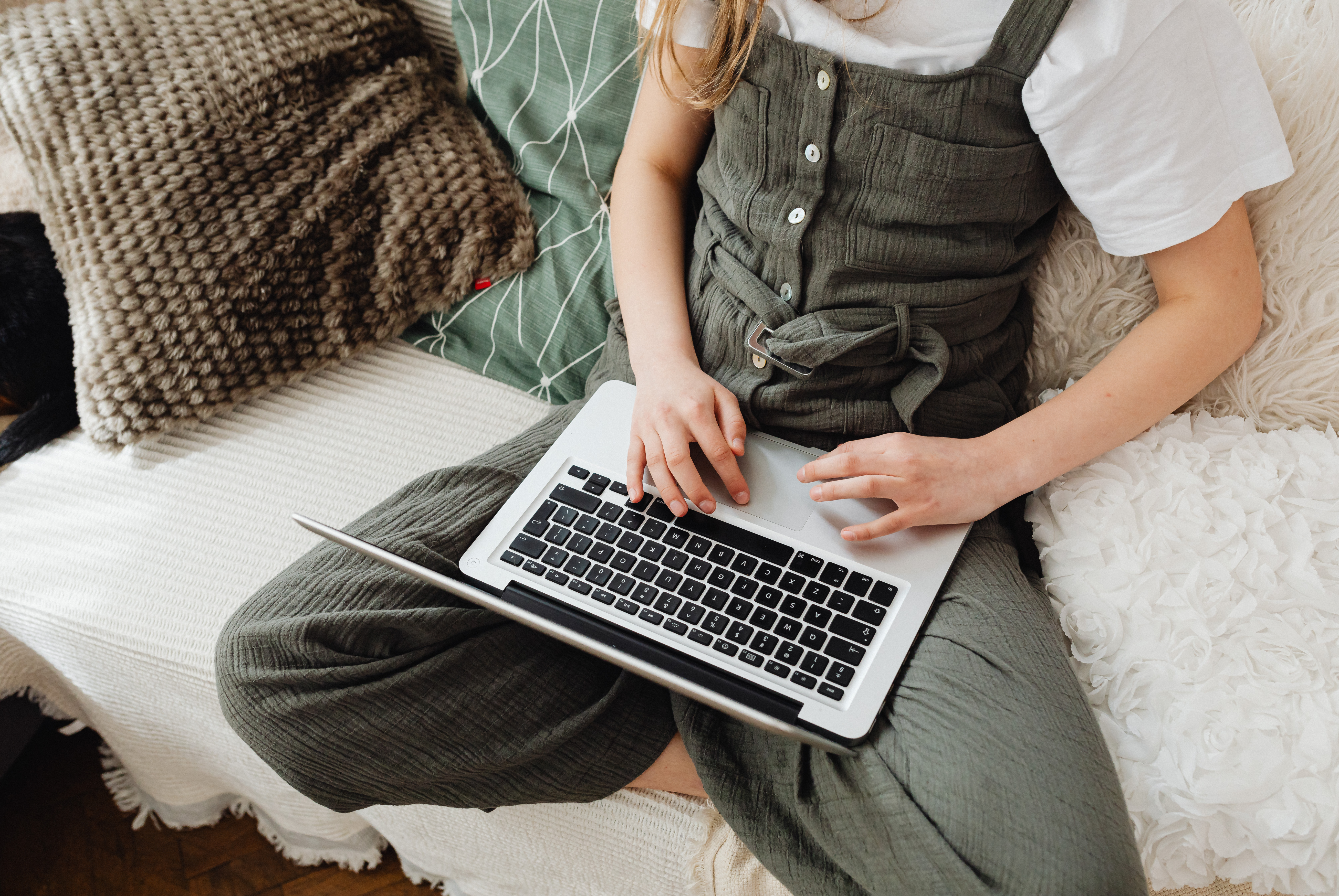 A young white woman sits on a couch working on a laptop, her face out of view